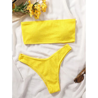 Bikini bandeau jaune effet froissé posé à plat sur un drap blanc, haut tubulaire et bas échancré près de fleurs jaunes