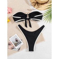 Bikini bandeau brésilien à anneau métallique noir à liseré crème posé à plat sur un tapis beige, bandeau noué devant