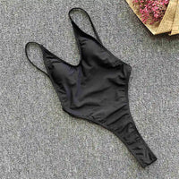 Maillot de bain une pièce noir à plat sur un tapis gris, bretelles fines et coupe string haute