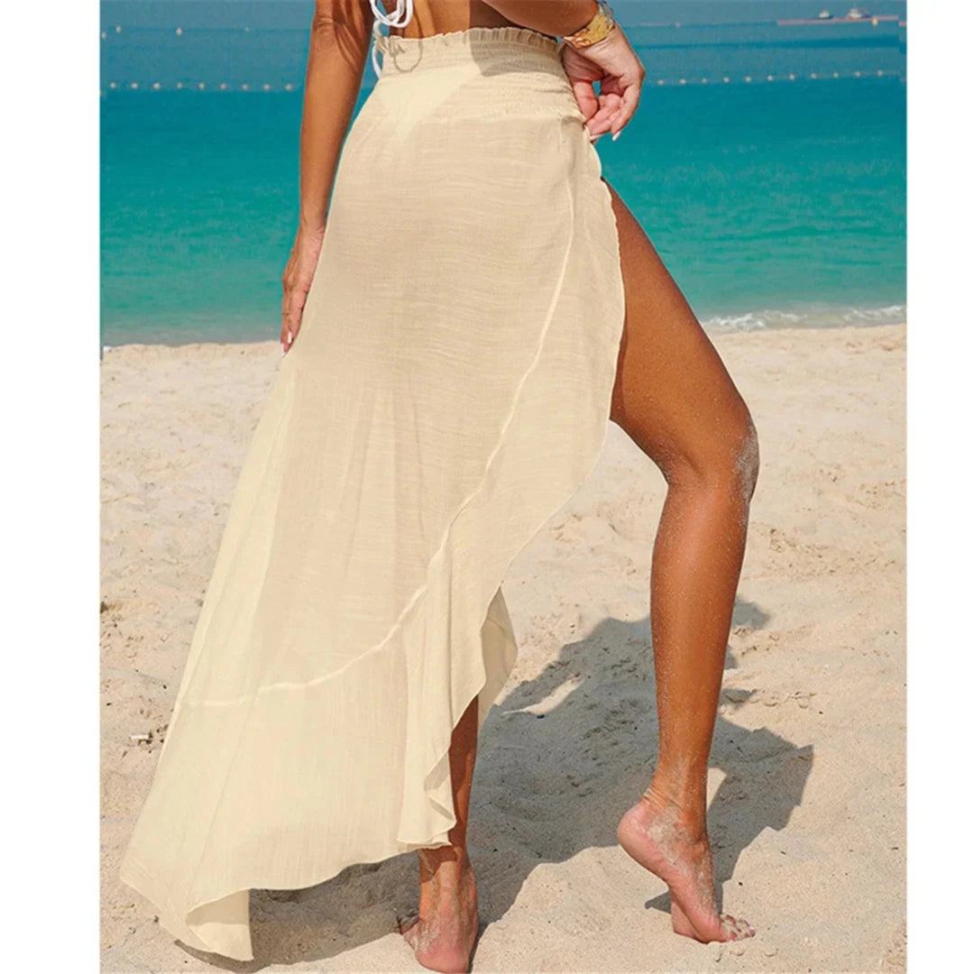 Jupe de plage beige vue de côté, bas asymétrique et fente haute, pieds nus sur le sable