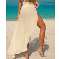 Jupe de plage beige vue de côté, bas asymétrique et fente haute, pieds nus sur le sable