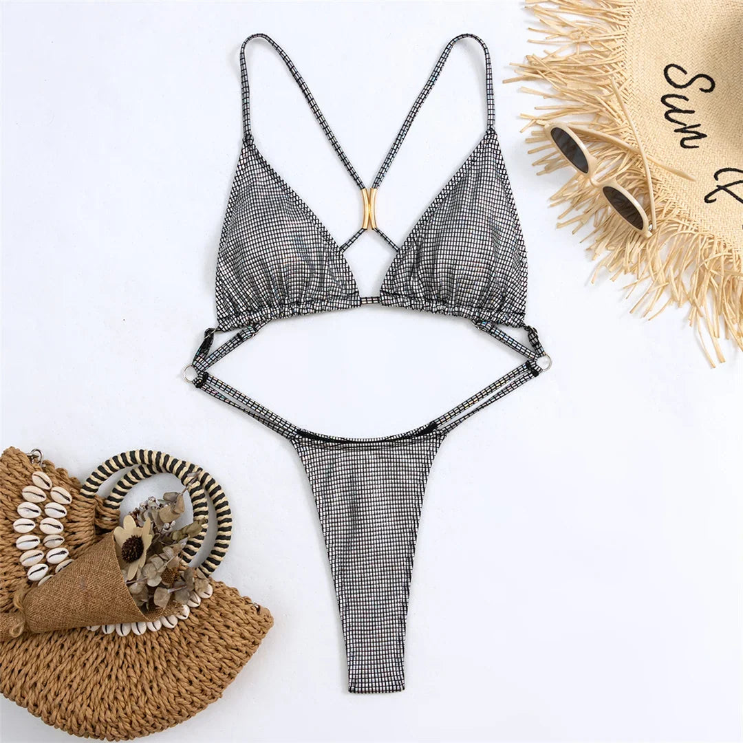 Maillot une pièce string argenté à plat sur fond blanc, bretelles croisées et anneau doré au centre, chapeau de paille à côté