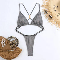 Maillot une pièce string argenté à plat sur fond blanc, bretelles croisées et anneau doré au centre, chapeau de paille à côté