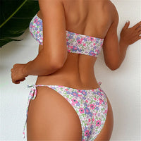 Bikini bandeau imprimé fleuri vu de dos, bas à nouer sur les côtés, sur fond de mur blanc et feuillage vert