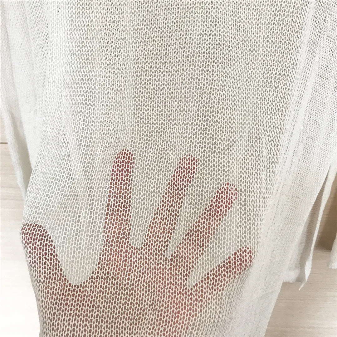 Gros plan sur le mesh blanc cassé d'une tunique de plage, la texture ajourée laisse apparaître une main dessous