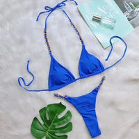 Bikini string tour de cou à anneau métallique froncé bleu posé à plat sur fourrure blanche, magazine et feuille verte