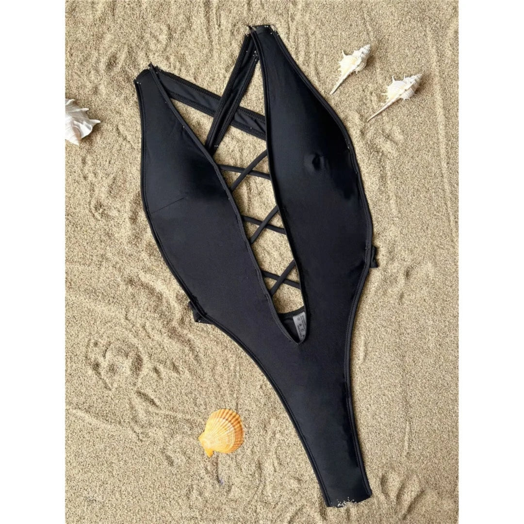 Maillot une pièce échancré noir étalé à plat sur du sable, décolleté en V et lanières croisées au centre, coquillages autour