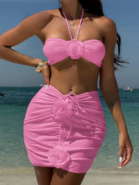 Maillot de bain trois pièces rose porté de face sur la plage, main sur la hanche, fleurs en relief sur la jupe froncée