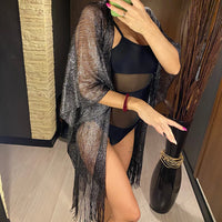 Kimono de plage à franges en maille brillante noir porté par-dessus un maillot noir, une main devant le visage