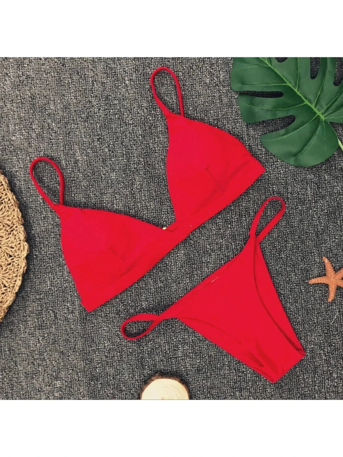 Bikini brésilien rembourré à taille basse rouge posé à plat sur tapis gris, bretelles fines et feuille de monstera