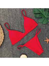 Bikini brésilien rembourré à taille basse rouge posé à plat sur tapis gris, bretelles fines et feuille de monstera