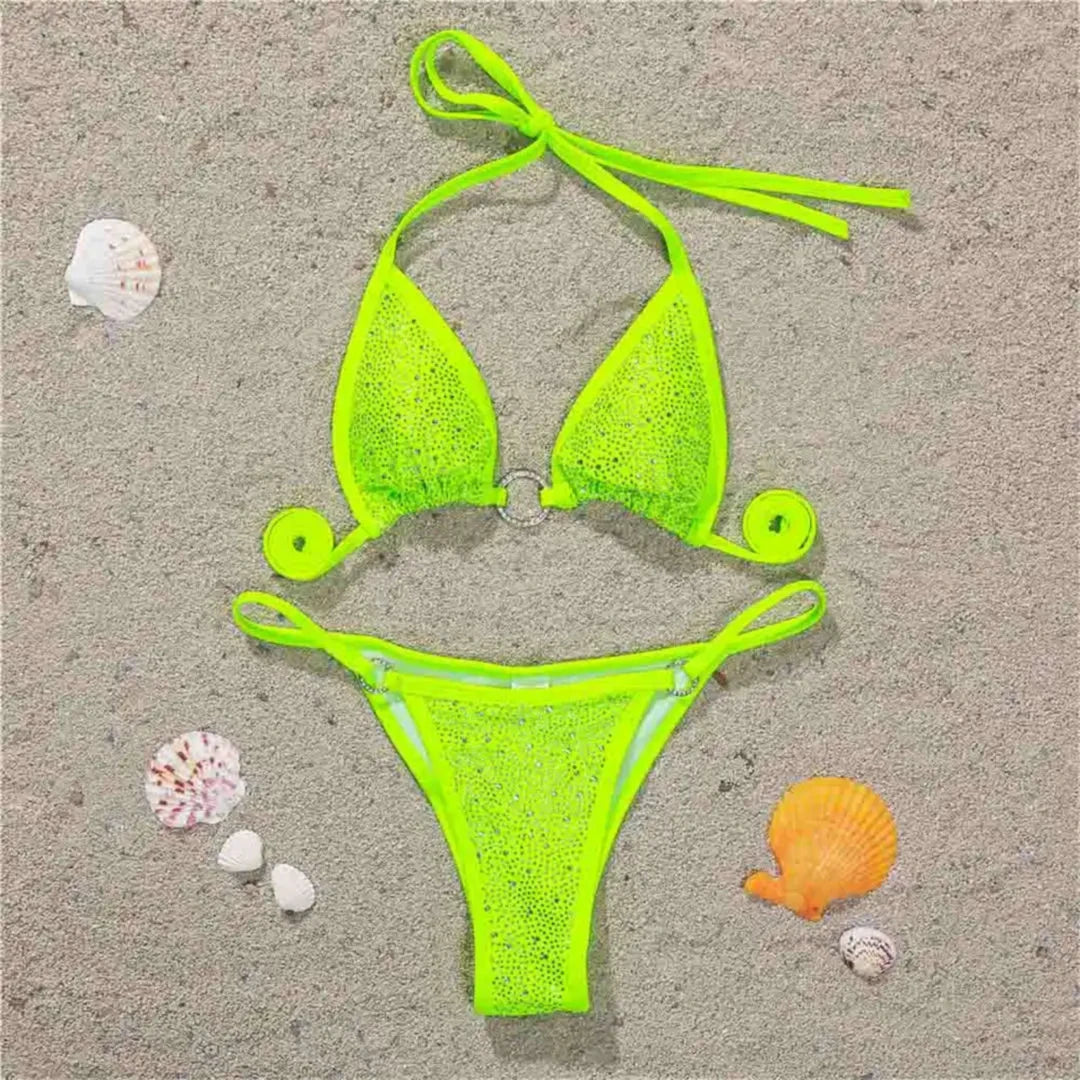 Bikini mini strass sans armatures taille basse vert fluo posé à plat sur du sable, strass et anneaux sur le haut