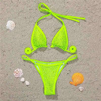 Bikini mini strass sans armatures taille basse vert fluo posé à plat sur du sable, strass et anneaux sur le haut