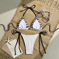 Bikini tour de cou blanc à liserés noirs posé à plat sur un tapis en jute, lunettes de soleil transparentes