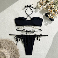 Bikini noir à liens latéraux posé à plat sur un tissu blanc, chapeau de paille et chaîne de coquillages