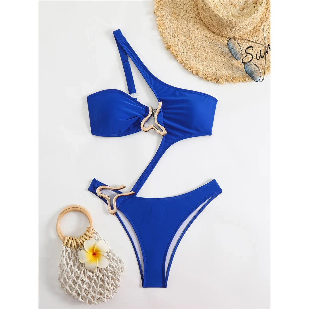 Maillot de bain une pièce asymétrique bleu posé à plat avec chapeau de paille, anneau doré au ventre et sac en filet