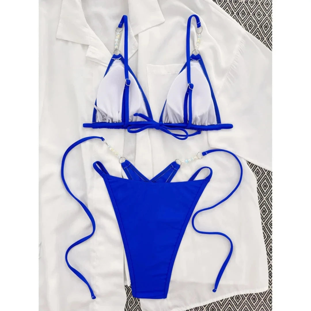 Bas de bikini bleu roi à lacets longs posé à plat sur une chemise blanche, vu de haut avec perles sur les côtés