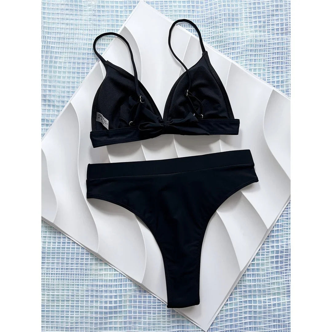 Bikini brésilien échancré taille haute noir vu à plat sur un motif blanc en vagues, haut croisé au centre et bretelles fines