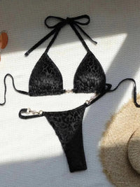 Bikini string micro à tour de cou imprimé léopard noir posé à plat sur un tissu blanc, anneau doré et liens longs