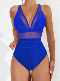 Maillot de bain une pièce décolleté en V en mesh bleu foncé porté de face par une femme devant un mur blanc, bandes transparentes