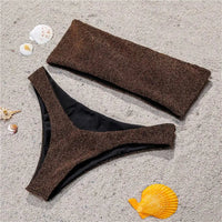 Bikini bandeau brillant pailleté marron étalé à plat sur un sable clair, à côté d'un coquillage orange et doublure noire