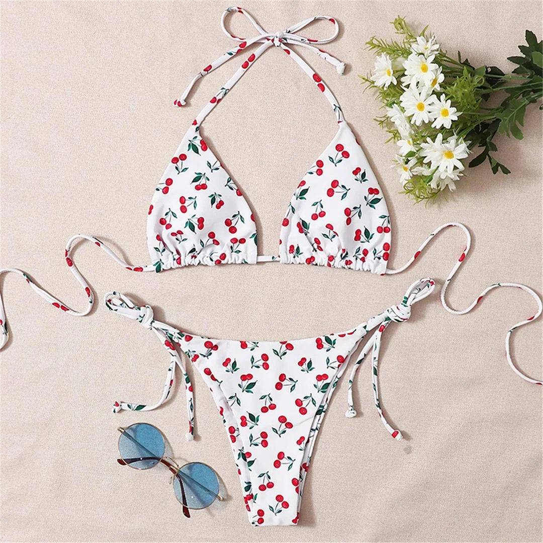 Bikini triangle tour de cou imprimé cerises rouges posé à plat sur un tissu beige, avec lunettes bleues et fleurs blanches