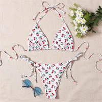 Bikini triangle tour de cou imprimé cerises rouges posé à plat sur un tissu beige, avec lunettes bleues et fleurs blanches