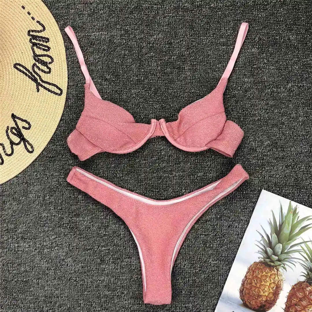 Bikini rose pailleté à armatures posé à plat sur un tapis gris, bas à liseré blanc, à côté d'un ananas