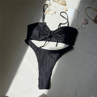 Bikini brésilien texturé froissé noir posé à plat sur un tapis, lumière du soleil et coquillages en arrière-plan