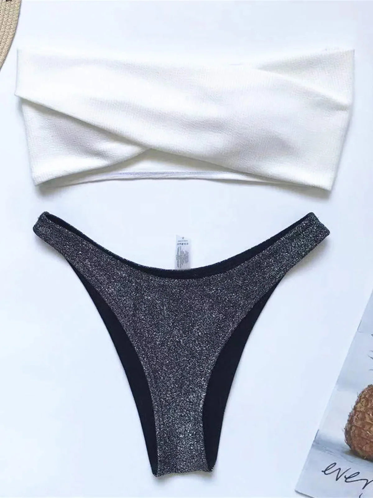 Bikini bandeau scintillant à paillettes noir posé à plat sur fond blanc, haut blanc torsadé et bas pailleté noir