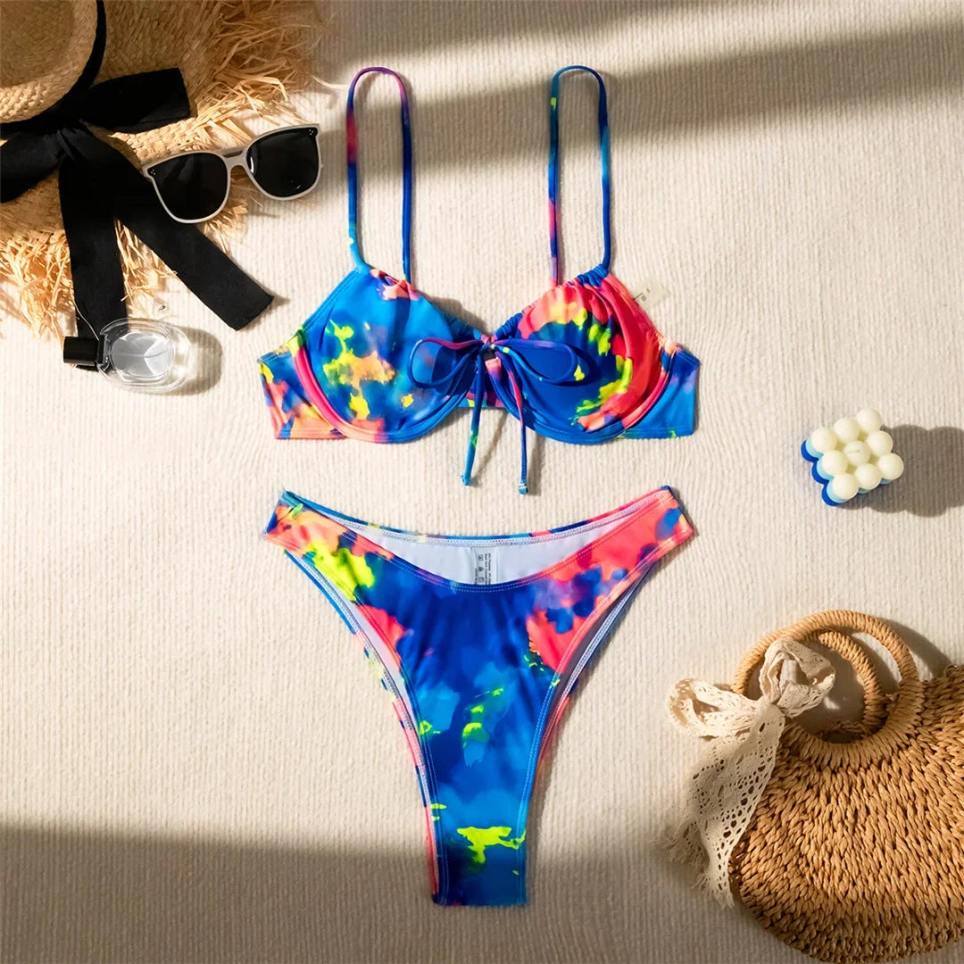 Bikini à armatures imprimé multicolore bleu, rose et jaune posé à plat sur du lin, chapeau de paille et lunettes à côté