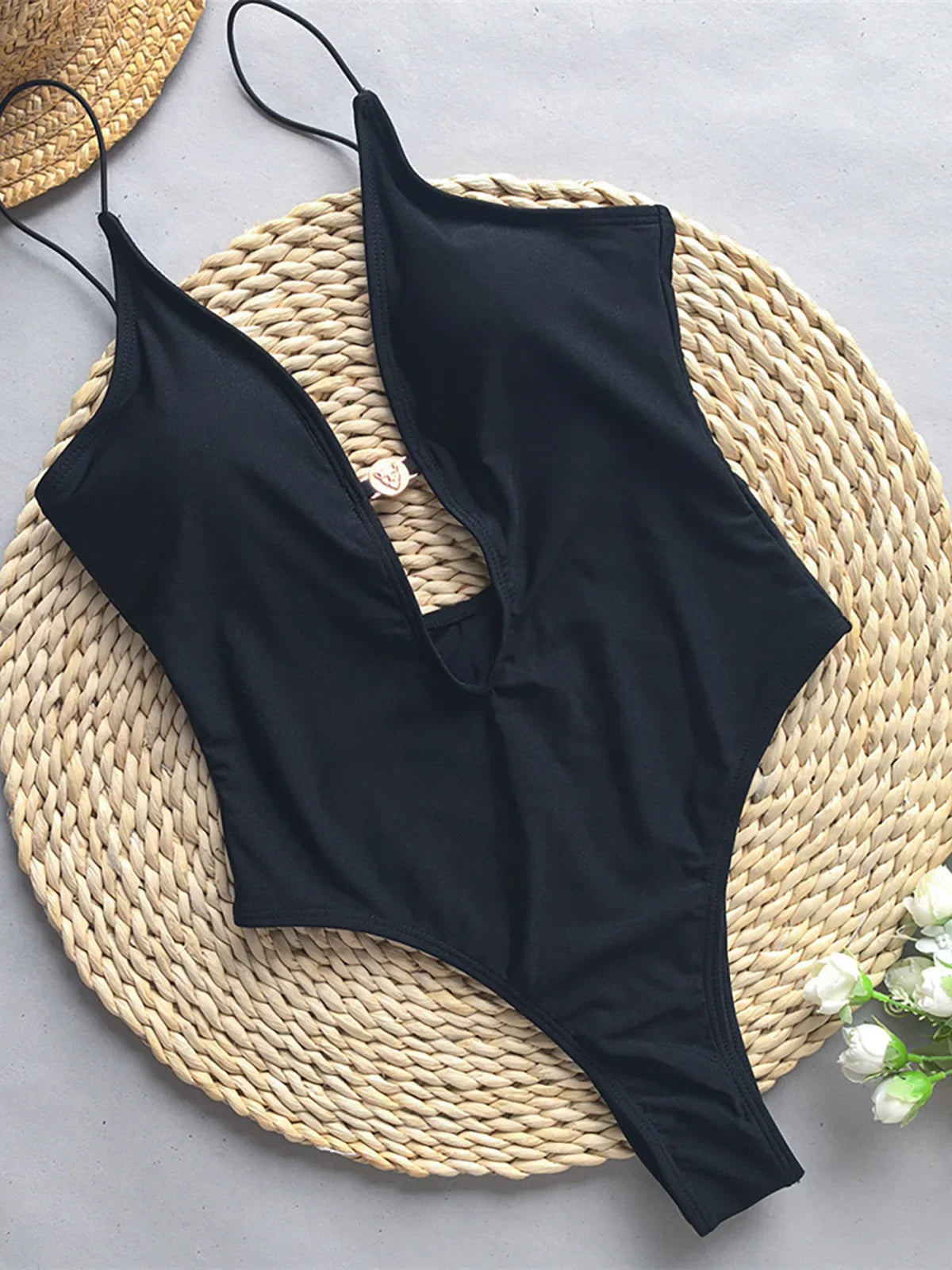 Maillot de bain une pièce échancré noir posé à plat sur un tapis en osier, décolleté plongeant et fines bretelles