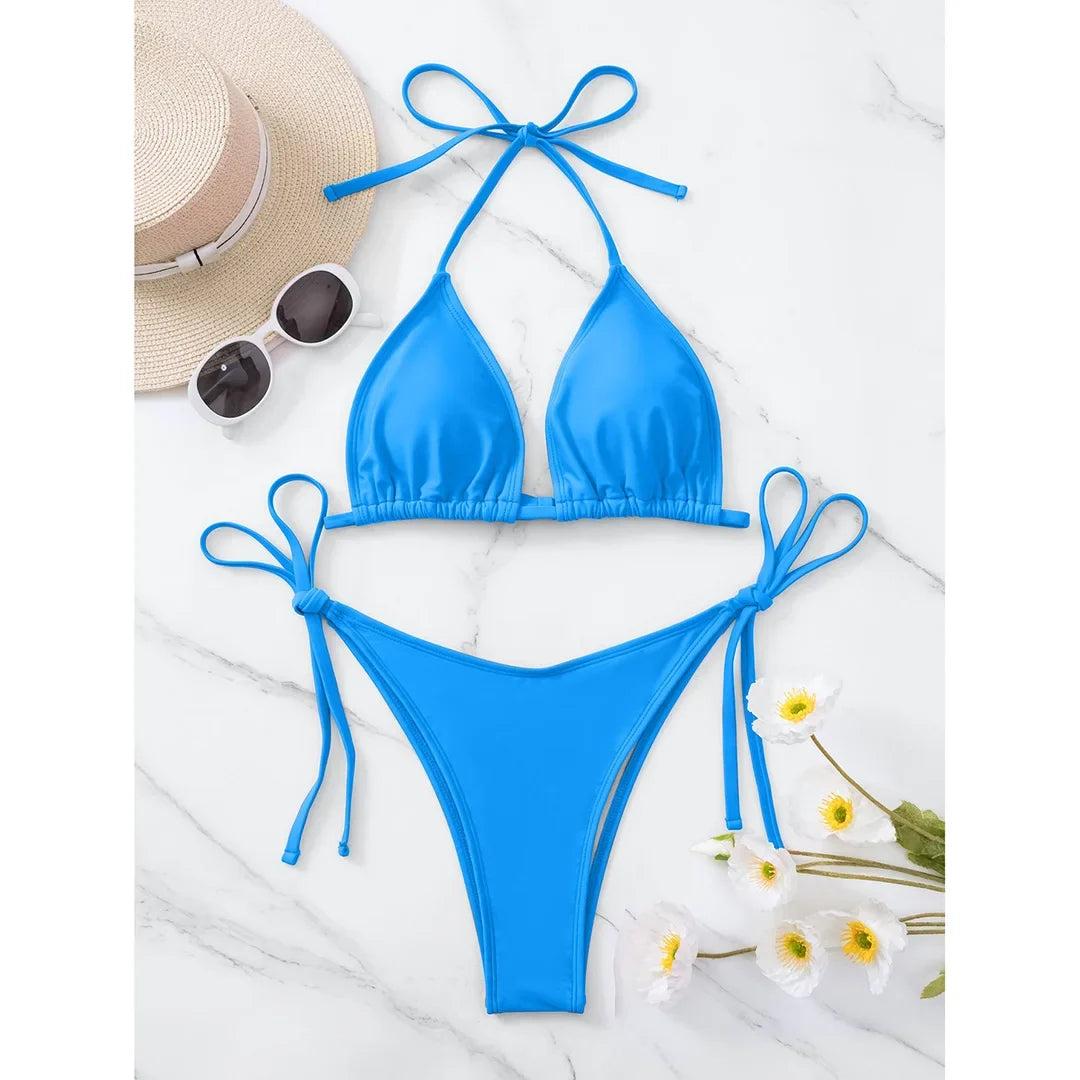 Bikini brésilien tour de cou rembourré uni bleu posé à plat sur marbre blanc avec chapeau et fleurs blanches