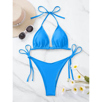Bikini brésilien tour de cou rembourré uni bleu posé à plat sur marbre blanc avec chapeau et fleurs blanches