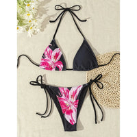 Bikini tour de cou sans armatures noir à grandes fleurs roses posé à plat sur une natte, près de marguerites et d'un sac rond