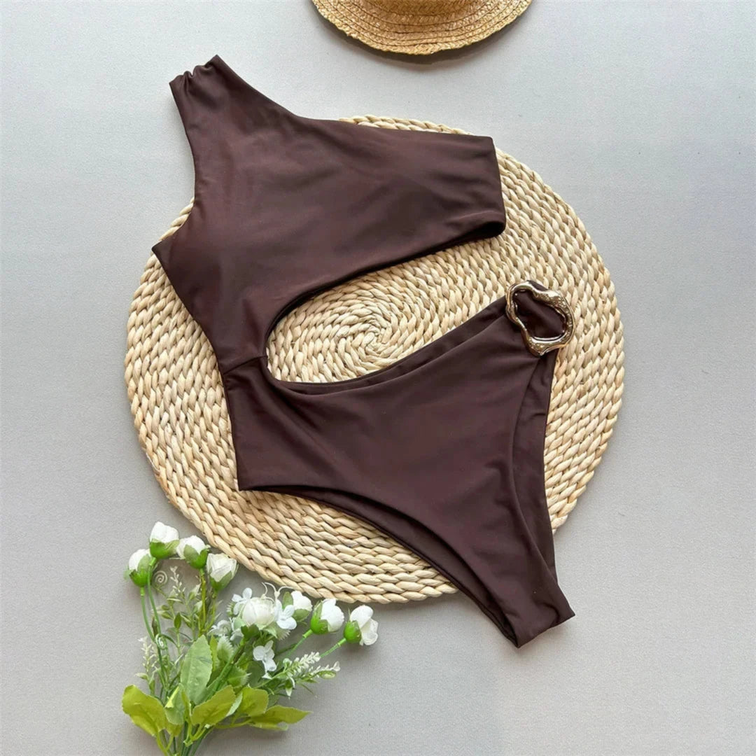 Maillot de bain une pièce asymétrique à anneau métal marron posé à plat sur un set rond en osier avec fleurs blanches