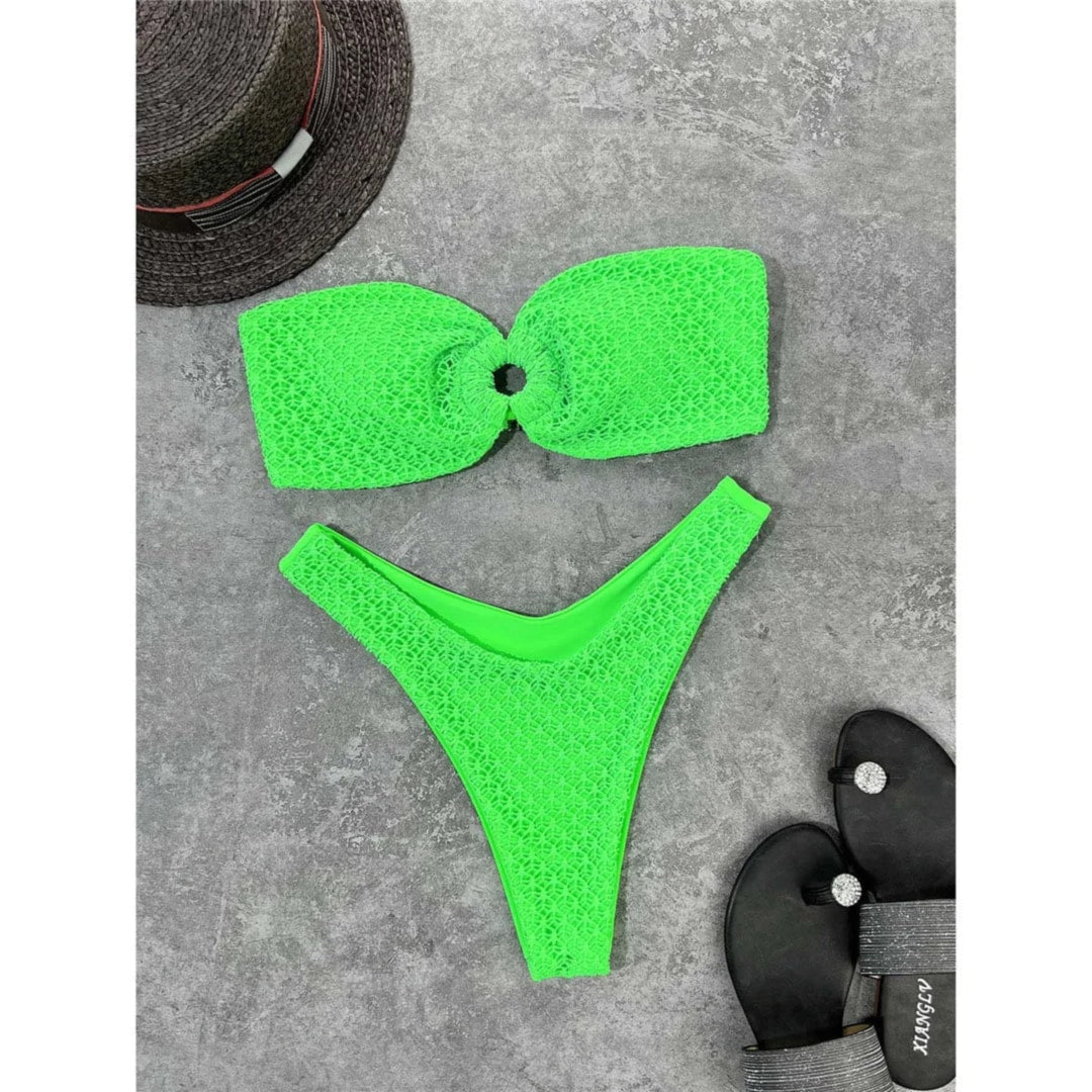 Bikini bandeau échancré vert fluo texturé posé à plat sur du béton gris, chapeau noir à côté