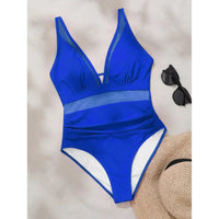 Maillot une pièce bleu foncé posé à plat sur fond clair, décolleté en V et bande de mesh à la taille, lunettes et chapeau en décor