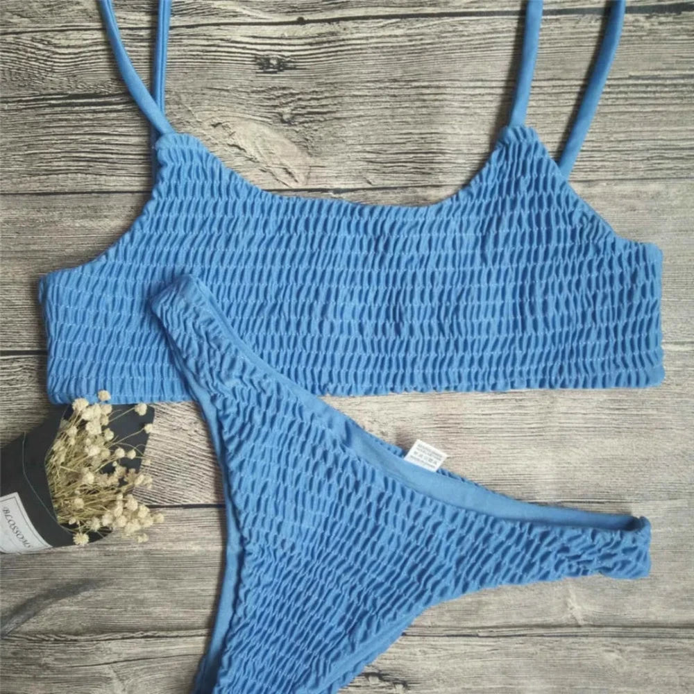 Bikini deux pièces texturé effet froissé bleu à plat sur une planche en bois, à côté d'une branche de fleurs séchées