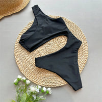 Maillot de bain une pièce asymétrique à anneau métal noir posé à plat sur un set rond en osier, chapeau de paille en haut