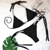 Bikini orné de strass brillants taille basse noir posé à plat sur un tapis blanc à côté d'une feuille verte