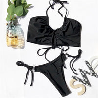 Bikini deux pièces noir à lacets posé à plat sur fond blanc, avec un vase en verre et des lettres décoratives