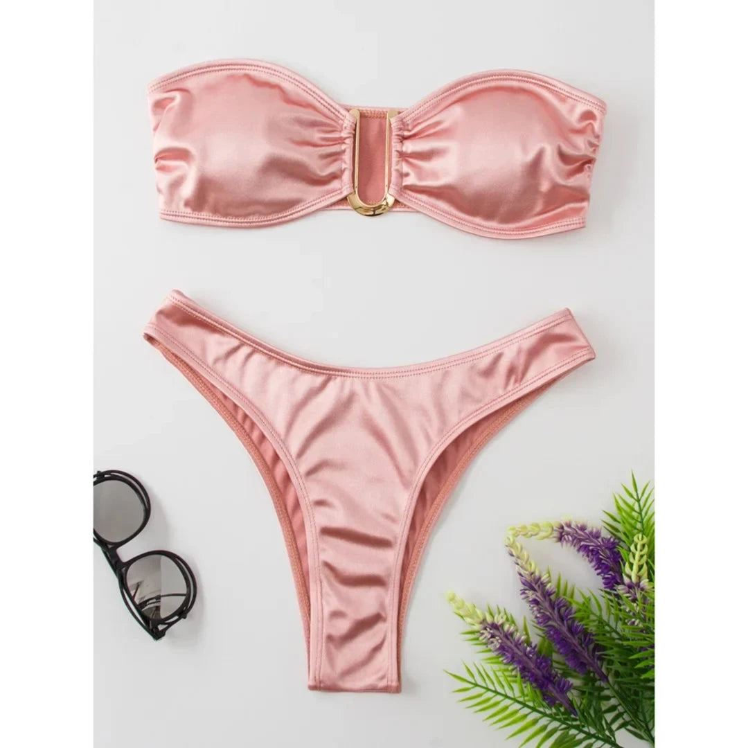 Bikini bandeau sans armatures taille basse rose posé à plat, lunettes de soleil et plante à côté, fond blanc