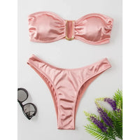 Bikini bandeau sans armatures taille basse rose posé à plat, lunettes de soleil et plante à côté, fond blanc