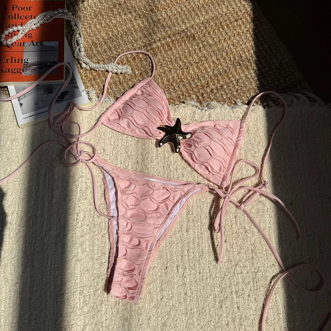 Bikini bandeau à lacets texturé rose poudré orné d'une étoile de mer, posé à plat sur un tissu au soleil