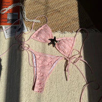 Bikini bandeau à lacets texturé rose poudré orné d'une étoile de mer, posé à plat sur un tissu au soleil