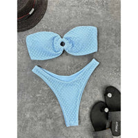 Bikini bandeau échancré bleu clair texturé posé à plat sur du béton gris, sandales noires à côté