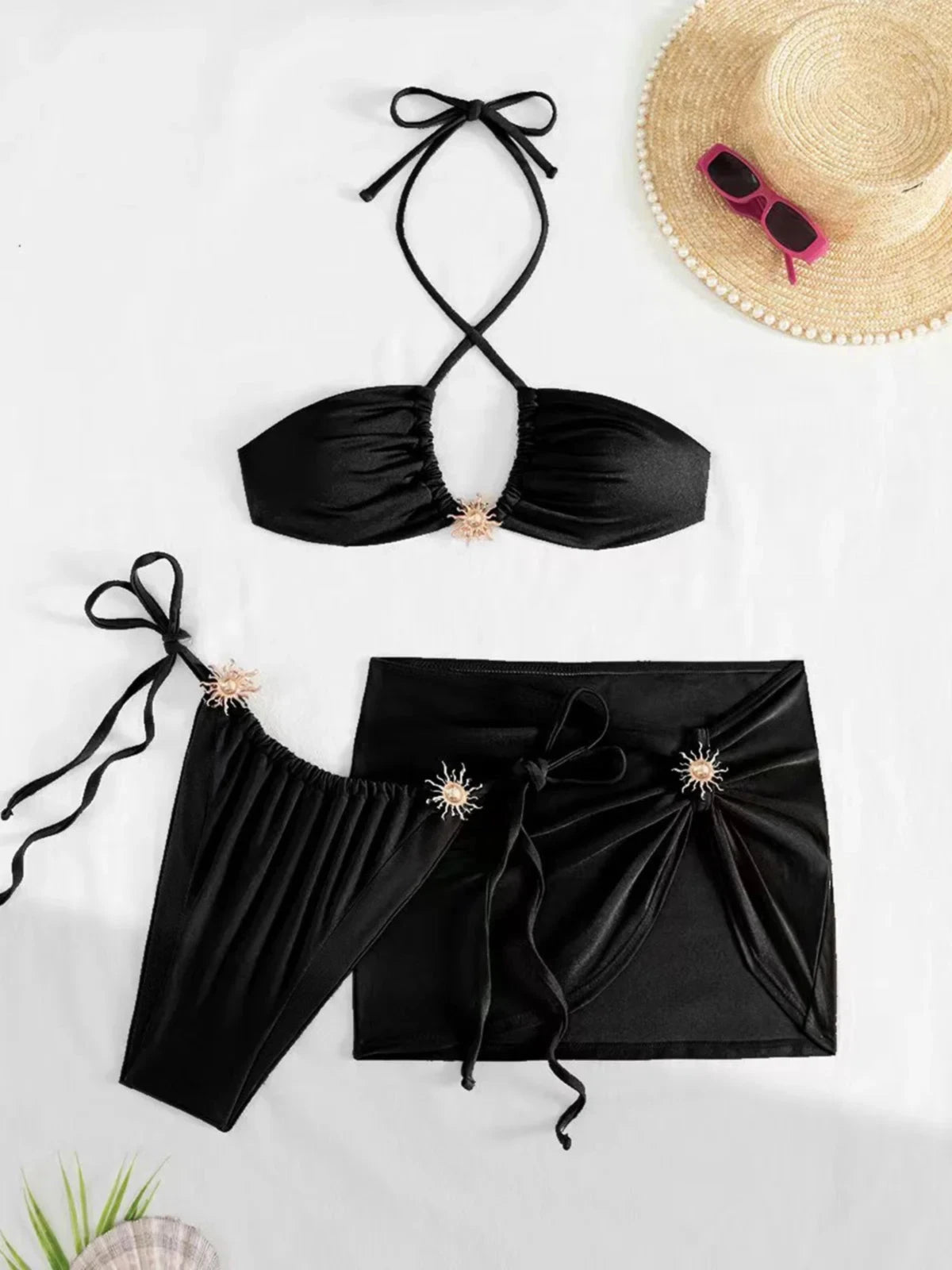 Maillot de bain trois pièces jupe halter à brides noir à plat sur fond blanc, avec chapeau de paille et lunettes de soleil posés à côté