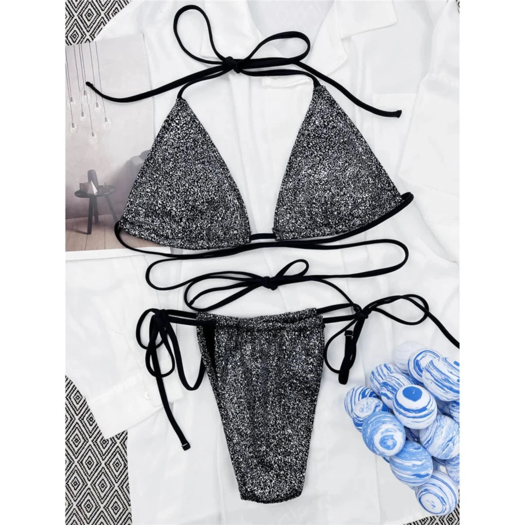 Bikini croisé argenté et noir à paillettes posé à plat sur un tissu blanc, haut triangle et bas à lacets latéraux