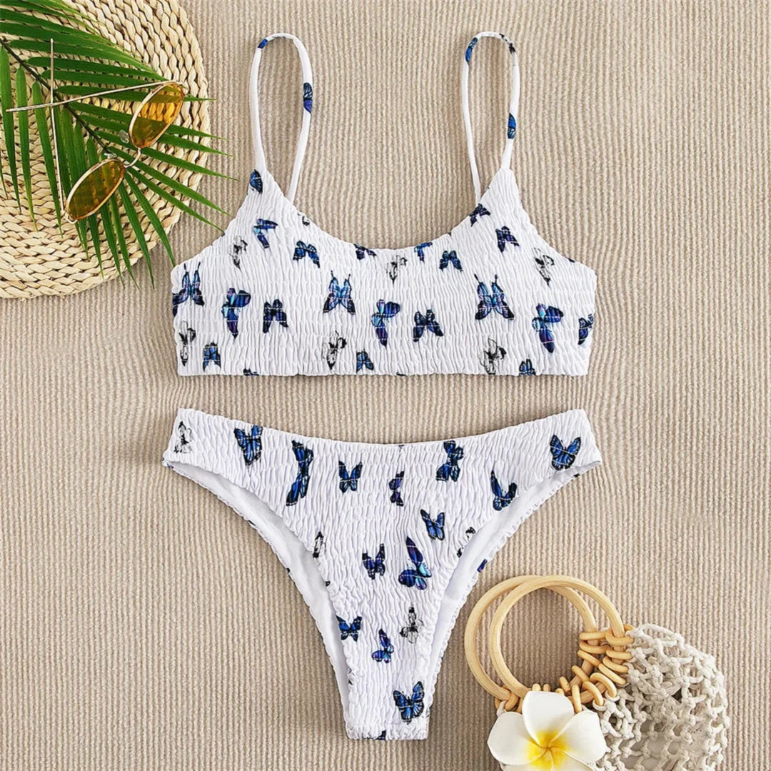 Bikini deux pièces texturé effet froissé imprimé papillons bleu à plat sur un tissu beige, avec chapeau de paille et fleur